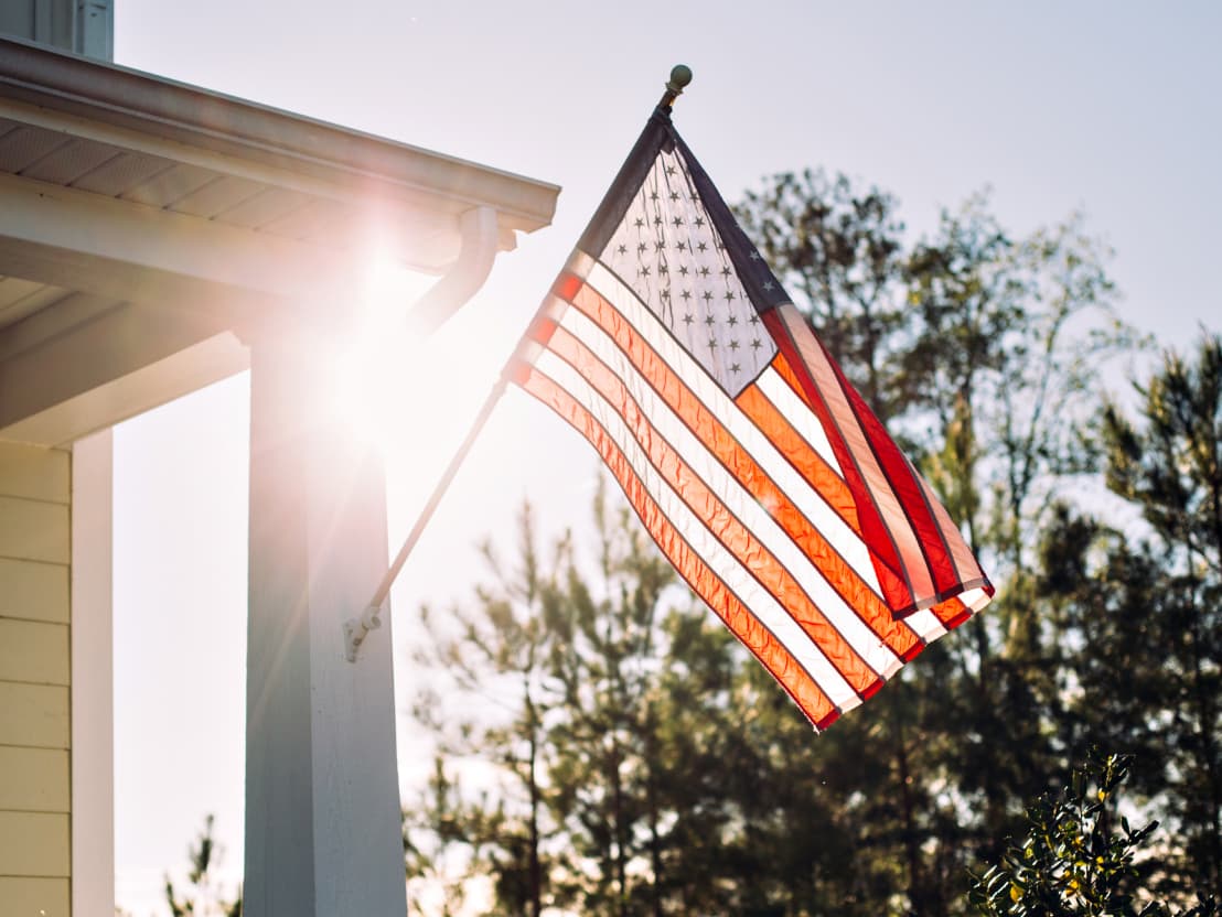 An American flag is displayed on a pole attached to the side of a building. The sun shines brightly through the trees in the background, creating a lens flare effect.